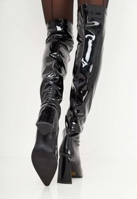Botas negras hasta el muslo, hechas de cuero sintético brillante, con tacón cuadrado y una silueta elegante que presenta una costura vertical en la parte posterior.