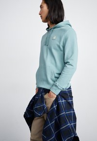 Felpa azzurra con tasca a marsupio e un piccolo logo sul petto, indossata sopra pantaloni beige, con una camicia a quadri blu e nera annodata in vita.