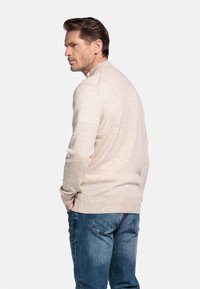 Man met kort bruin haar, gekleed in een beige sweater en blauwe jeans, staand met handen in de zakken, kijkend naar de zijkant op een witte achtergrond.