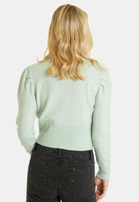 Cardigan leggero a maglia di colore verde menta, con orlo a coste e maniche leggermente a sbuffo. Indossato con jeans neri caratterizzati da decorazioni metalliche.