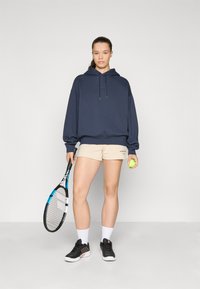 Felpa navy con cappuccio e coulisse, pantaloni corti beige, scarpe da ginnastica nere, tiene in mano una racchetta da tennis e una pallina da tennis verde.