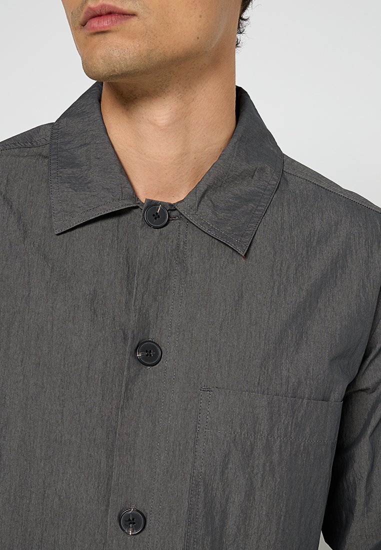 Szary koszula z guzikami z wystrzałowym kołnierzykiem, wykonana z teksturowanej tkaniny. Posiada czarne guziki i kieszeń na piersi.