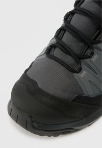 Close-up de um sapatilha atlética preta e cinza escuro com tecido texturizado, atacadores pretos e sola de borracha resistente em fundo branco.