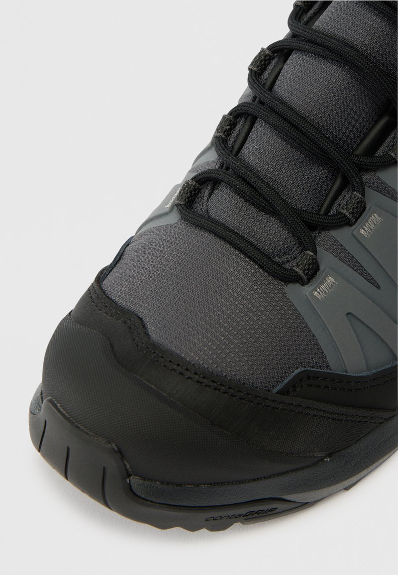 Nahaufnahme eines schwarzen und dunkelgrauen Sport Schuhs mit strukturiertem Stoff, schwarzen Schnürsenkeln und robusten Gummisohlen auf einem weißen Hintergrund.