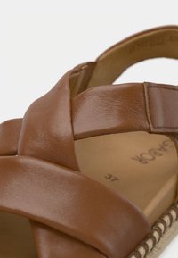 Sandale en cuir marron avec un design à brides croisées, une surface texturée et une semelle en fibre naturelle. Comprend une bride de cheville sécurisée.