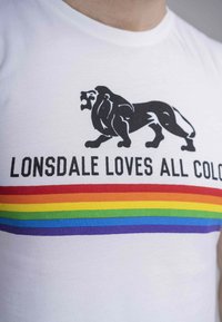 Hvid bomulds t-shirt med et sort løvegrafik og teksten "LONSDALE ELSKER ALLE FARVER" over et vandret regnbuestribemønster.