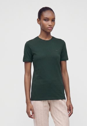 Femme à la peau foncée portant une chemise à manches courtes vert foncé et un pantalon beige clair à motifs, debout devant un fond clair uni.