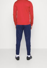 Chemise rouge à manches longues assortie avec un pantalon de sport bleu marine. La chemise a une coupe ajustée ; le pantalon est fuselé. Des chaussures de sport noires sont portées.