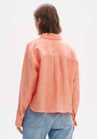 Perzikkleurig shirt met een relaxte pasvorm, lange mouwen en een kraag. Beschikt over een plooi aan de achterkant en knoopmanchetten. Gecombineerd met lichtblauwe spijkerbroek.