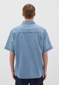 Camisa de denimp de manga corta de color azul claro con cuello, costuras visibles y un corte relajado. La textura parece suave y ligeramente desgastada.