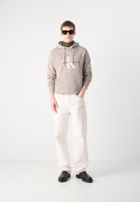 Felpa con cappuccio in beige chiaro con logo, abbinata a jeans bianchi a gamba dritta e scarpe slip-on nere. Modello che indossa occhiali da sole neri.