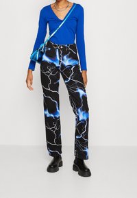 Person med en blå långärmad topp, svarta byxor med blå blixttryck, svarta stövlar, silverkedjehalsband och en metallisk blå axelväska.