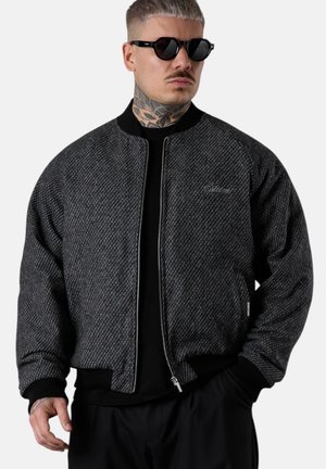 Mann mit Hals-Tattoo und kurzem Haar, der schwarze Sonnenbrillen und eine dunkelgraue Bomberjacke mit Muster über schwarzer Kleidung trägt, steht mit den Händen in den Taschen.