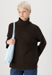 Brūna pārāk liela adīta turtlenecka džemperis ar ribota akcentiem, savienots ar gaiši zilu somu. Modelis valkā zilus džinsus un viņai ir īsi mati.