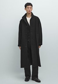 Cappotto lungo nero con colletto a punta, chiusura con bottoni e tasche laterali. Indossato sopra un maglione nero e pantaloni, presenta un design semplice e pulito.