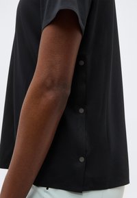 Πλάγια όψη ενός ατόμου που φοράει μαύρο πουκάμισο με κοντά μανίκια και τρία εμφανή κουμπιά στο πλαϊνό ραφτό.