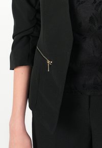 Svart blazer med en strukturerad design, med en dekorativ dragkedjeficka på sidan. Materialet verkar slätt med en mjuk textur.