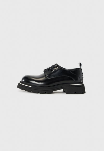Chaussure en cuir noir avec une finition brillante, bout rond, laçage à six œillets et une semelle noire épaisse et texturée avec des accents blancs.