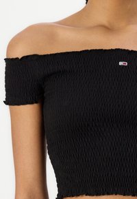 Top nero senza spalle realizzato in tessuto smock a trama; presenta maniche corte e un piccolo logo in bianco, rosso e blu.