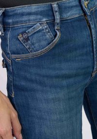 Jean en denim bleu à taille haute, avec une poche avant, une braguette zippée et des coutures dorées contrastantes. Texture lisse, design ajusté.
