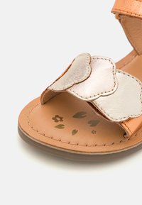 Sandalo in pelle color cuoio con cinturino metallico a forma di conchiglia, soletta imbottita con stampe floreali e suola in gomma marrone chiaro.