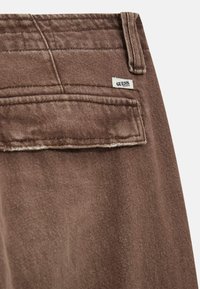 Bruine denimstof met een achterzak en een klein "GUESS" label. De textuur lijkt zacht met zichtbare stiksel-details.