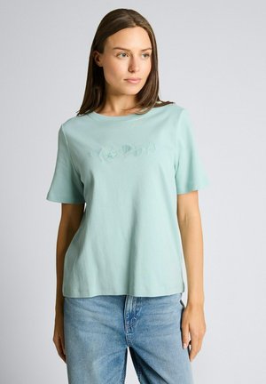 Frau mit glattem braunem Haar, die ein hellgrünes Kurzarm-Oberteil mit dezenten Blumenstickereien und hellblaue Jeans trägt, steht vor einem schlichten Hintergrund.