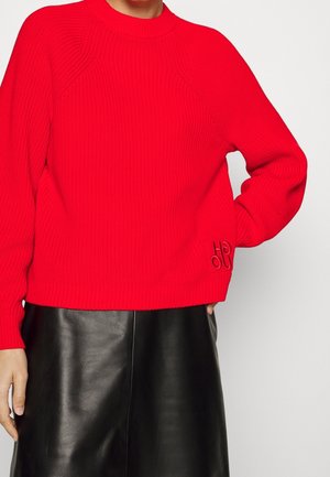 Personne portant un pull en laine rouge vif à manches longues et une jupe en cuir noir, main posée sur la hanche, sur un fond uni.