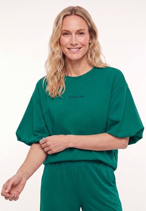 Lachende vrouw met golvend blond haar draagt losse groene top en broek en past haar mouw aan tegen een lichte effen achtergrond.