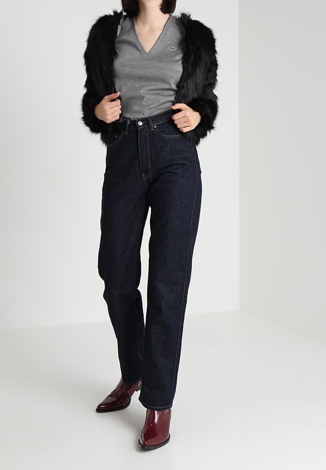 Femme portant un t-shirt gris à col en V, une veste en fausse fourrure noire, un jean taille haute foncé et des bottines bordeaux, debout.