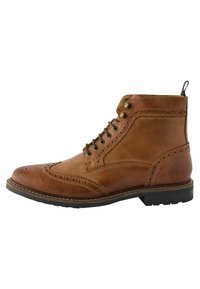 Botín de cuero marrón con detalles de brogue, punta redondeada, cordones en la parte frontal y suela de goma de contraste. Presenta una lengüeta en el talón.