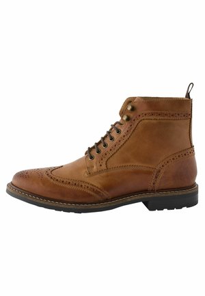 Botín de cuero marrón con detalles de brogue, punta redondeada, cordones en la parte frontal y suela de goma de contraste. Presenta una lengüeta en el talón.