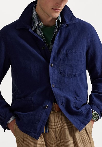 Navy blue linen jacket with a spread collar, a left chest pocket, and two front pockets, layered over a plaid shirt and brown plaid trousers.