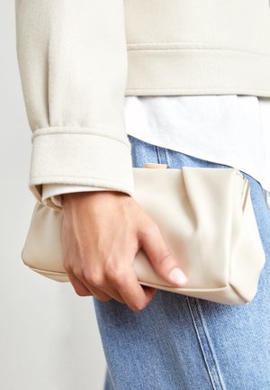 Ręka trzymająca miękki beżowy kopertówkę, ubrana w jasnoniebieskie dżinsy i kremową rękawicę kurtki.