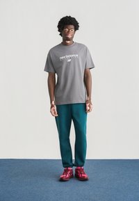 Γκρι T-shirt με λογότυπο «new balance», τιρκουάζ παντελόνι και ροζ αθλητικά παπούτσια. Το μοντέλο στέκεται μπροστά σε απλό φόντο, παρουσιάζοντας αθλητική ένδυση.