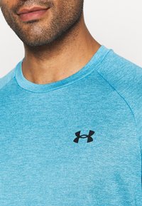 Närbild av en mans nedre ansikte och hals iklädd en ljusblå Under Armour-tröja med en svart logotyp på bröstet.