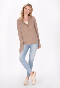 Långärmad, ljusbrun henley-tröja med texturerad yta och knappslå, parat med blå skinnyjeans och beige sneakers.