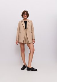Béžový dvouřadý blazer přes černý top, doplněný béžovou sukní a černými mokasínami. Blazer má zaoblené klopy a knoflíky.