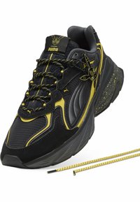Baskets Puma noires et jaunes avec semelle épaisse, talon taché de jaune, détails Pikachu sur la languette et le côté, et lacets à motif jaune et noir.