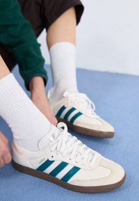 White sneakers with teal three-stripe design, featuring a smooth leather upper and brown rubber sole, worn with white ribbed socks.