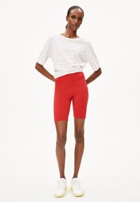 Rode fietsbroek gemaakt van rekbare stof, gecombineerd met een losvallend wit gestreept shirt. Model staat in witte sneakers.