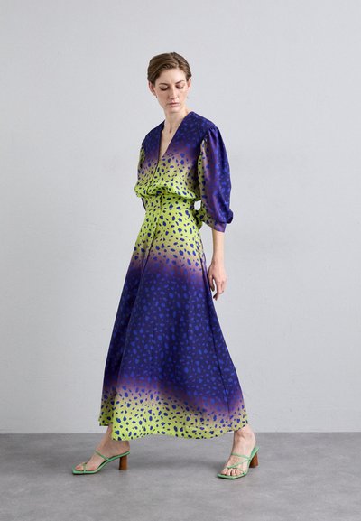 Maxi jurk in blauw en groen ombré, met een V-hals, ballonslouwen en een getailleerde taille. Acцентuated met een stippenpatroon en gedragen met groene sandalen.