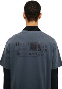 Grå kortærmet t-shirt med et sort grafik på ryggen, der indeholder forskellige tekster og symboler, sat mod en glat stofstruktur.