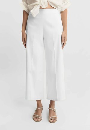 Donna che indossa pantaloni bianchi a vita alta, larghi e corti abbinati a sandali piatti color nudo con cinturini e camicetta bianca annodata.