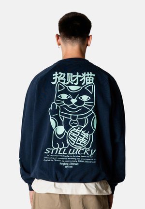 Mand iført marineblå sweatshirt med stort lyseblåt heldig katte-motiv og kinesiske tegn på ryggen, beige bukser og hvid skjorte under.