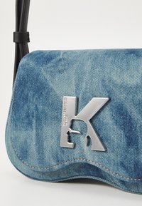 Džínová crossbody taška v světle modré barvě se stříbrným logem 'K'. Obsahuje zakřivený klapkový uzávěr a kontrastní oranžové švy.