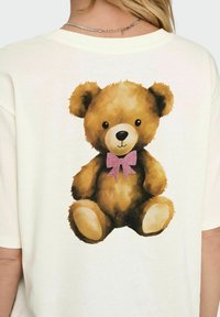 Crème-kleurig t-shirt met een grote, cartoonachtige bruine teddybeer met een roze glitter strik, geplaatst op de rug. Zachte stofstructuur.