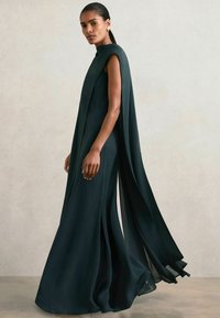 Langes, fließendes, dunkelgrünes Kleid aus leichtem Stoff mit hohem Kragen und kapellenartigen Ärmeln, das seitliche Schlitze und minimales Zubehör aufweist.