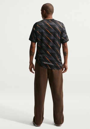 Muž stojaci chrbtom, oblečený v čiernej krátkorukávovej košeli s oranžovými a sivými diagonálnymi pruhmi, v hnedom nohaviciach a bielych teniskách.