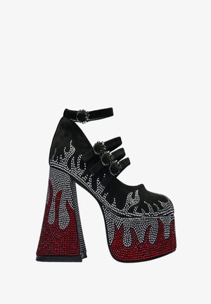 Tacón plataforma negro con correas en el tobillo y hebillas, decorado con pedrería plateada y roja que forma patrones de llamas.